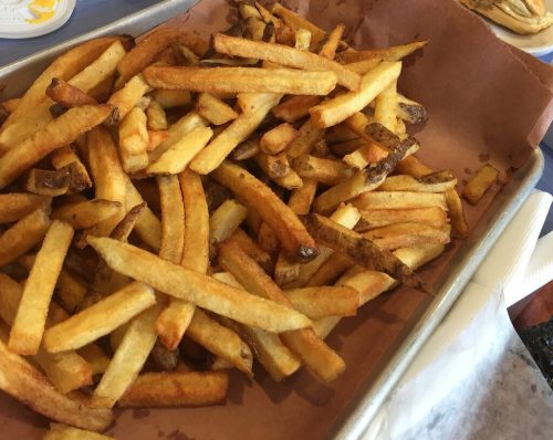 A tray of golden, crispy French fries on a table, with a small cup of sauce and a partially eaten plate in the background.
