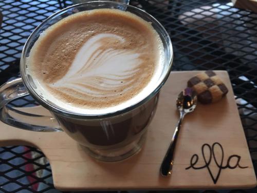 A cup of latte with a heart-shaped foam design, served on a wooden board with a small cookie and a spoon.