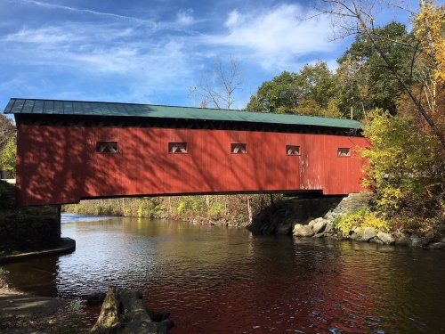 A red covered bridge spans a calm river, surrounded by trees and a clear blue sky.