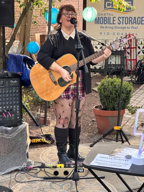 A musician with glasses sings and plays guitar outdoors, wearing a black jacket, plaid skirt, and fishnet tights.