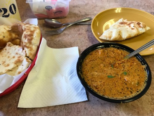 A bowl of creamy curry with a spoon, accompanied by naan bread and a plate with a stuffed pastry on a table.