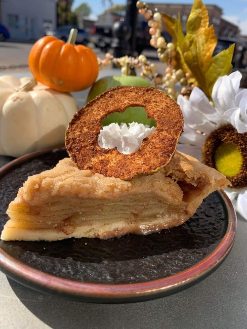 A slice of apple pie topped with a dried apple slice, surrounded by decorative pumpkins and autumn foliage.