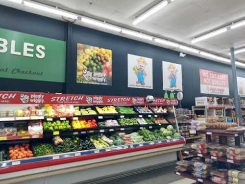 A grocery store produce section with various fruits and vegetables, featuring promotional signs and a checkout area.