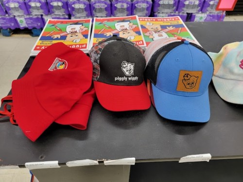 Three hats displayed on a counter: a red cap, a black and red cap with a logo, and a blue cap with a pig logo.