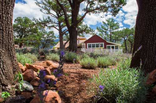 A charming red house surrounded by trees and purple flowers under a blue sky with fluffy clouds.