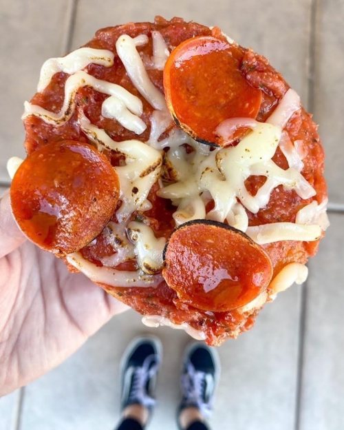 A hand holding a small pizza topped with pepperoni and melted cheese, with sneakers visible in the background.