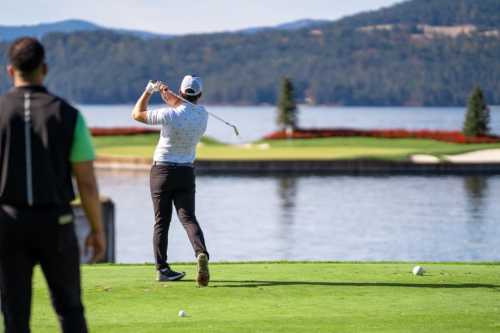 A golfer swings a club on a sunny day, with a lake and mountains in the background. Another person stands nearby.