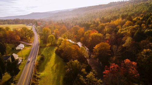 Aerial view of a winding road alongside a river, surrounded by vibrant autumn foliage and rolling hills.