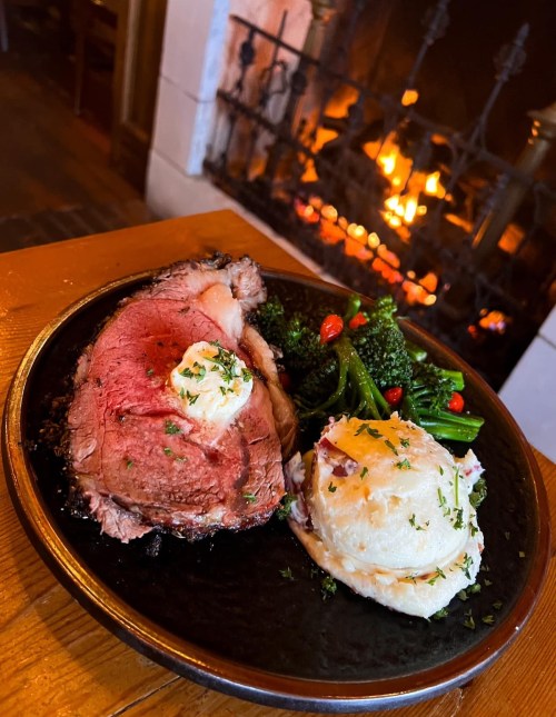A plate of prime rib with butter, mashed potatoes, and broccoli, set against a cozy fireplace backdrop.