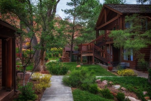 A serene garden path leads between rustic wooden cabins surrounded by lush greenery and colorful flowers.