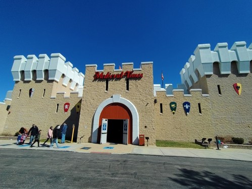 Entrance to Medieval Times, featuring a castle-like facade with colorful shields and a clear blue sky.