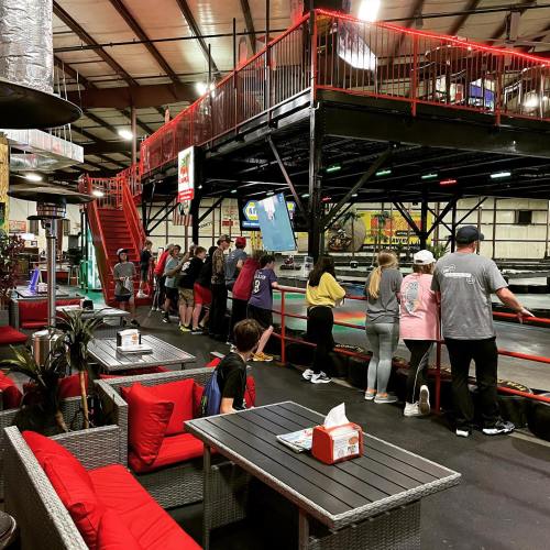 A lively indoor go-kart track with people waiting in line, surrounded by seating areas and colorful lighting.