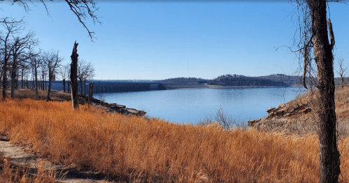 A serene lake surrounded by dry grass and trees, with a dam visible in the distance under a clear blue sky.