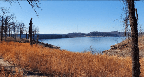 A serene lake scene with dry grass in the foreground and a dam in the background under a clear blue sky.