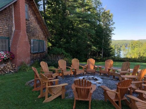 A cozy outdoor seating area with wooden chairs arranged around a fire pit, surrounded by trees and a lake in the background.