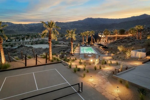 A desert landscape at sunset featuring a pool, palm trees, and a tennis court surrounded by mountains.