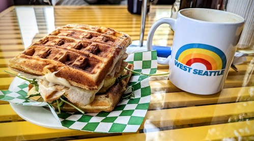 A waffle sandwich with chicken and greens next to a coffee mug featuring a rainbow design and "West Seattle" text.