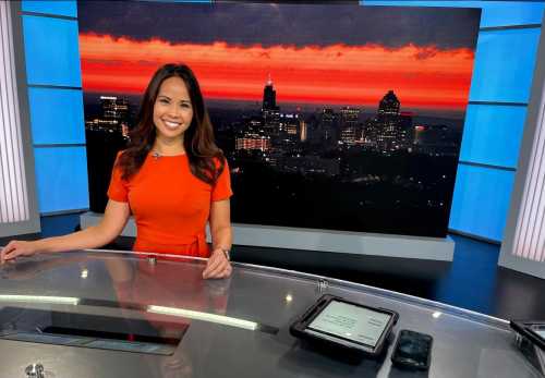 A news anchor in an orange dress smiles at the camera, with a vibrant sunset and city skyline in the background.