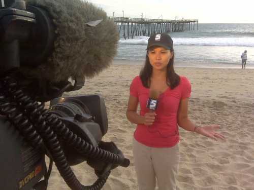 A reporter in a pink shirt and cap stands on the beach, speaking into a microphone with a camera in the foreground.