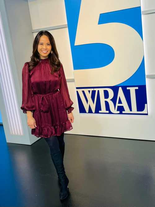 A woman in a burgundy dress and black boots stands in front of a large WRAL 5 sign in a studio setting.