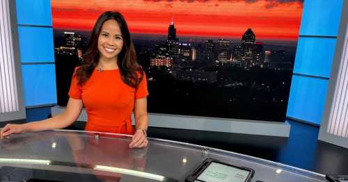 A news anchor in an orange dress smiles at the camera, with a vibrant sunset and city skyline in the background.