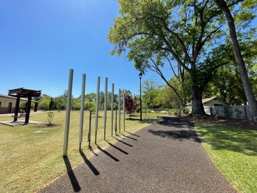 A sunny park path lined with tall poles, surrounded by green grass and trees under a clear blue sky.