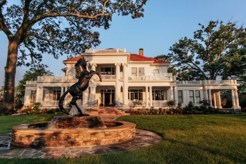 A grand white mansion with a horse statue in front, surrounded by lush green grass and trees under a clear sky.