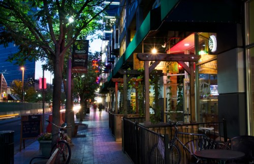 A quiet city street at night, featuring outdoor seating, trees, and illuminated storefronts.