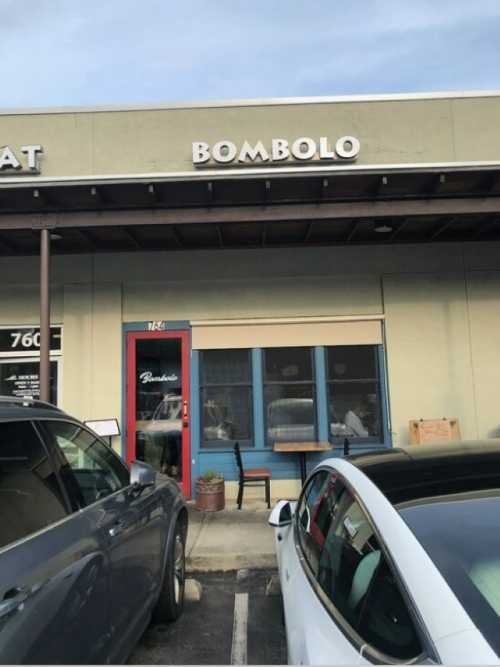 Exterior of a restaurant named "Bombolo," featuring a blue door and a small table outside. Cars parked in front.