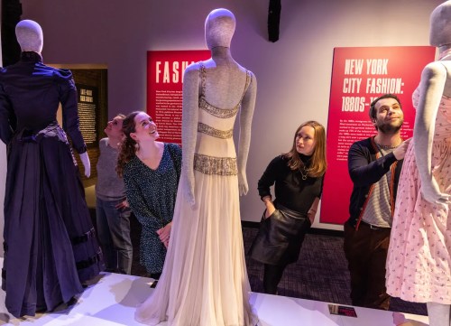 Visitors admire vintage dresses displayed on mannequins at a fashion exhibit. Bright pink informational panels in the background.