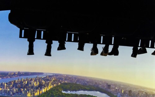 Silhouetted feet dangling from a ride, overlooking a cityscape with a river and green park at sunset.