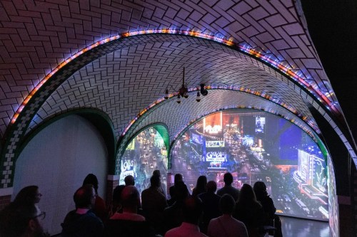 A group of people watching a vibrant, illuminated projection of a cityscape in an arched, tiled space.