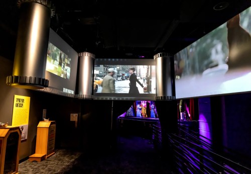 Three large screens display scenes from a film in a dimly lit exhibition space with colorful lighting.