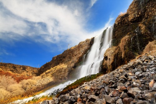 A cascading waterfall flows down rocky cliffs under a blue sky with wispy clouds.