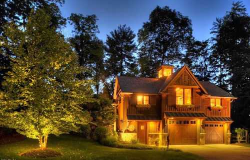 A cozy wooden house illuminated at dusk, surrounded by trees and a well-lit yard.