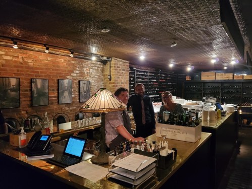 A cozy bar with brick walls, three staff members smiling behind the counter, and shelves filled with wine bottles.