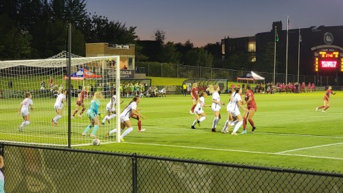 A soccer match in progress, with players from both teams near the goal, under stadium lights at dusk.