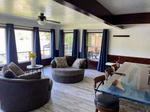 Cozy living area with two circular sofas, a dining table, and large windows with blue curtains, overlooking a scenic view.