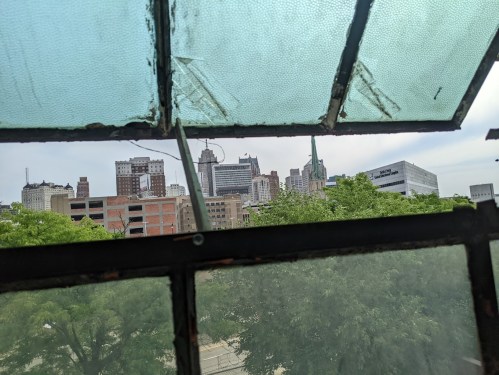 View of a city skyline through a partially open, weathered window, framed by green trees and a cloudy sky.