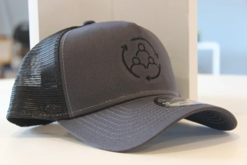 A gray baseball cap with a black mesh back, featuring a circular logo with three stylized figures.