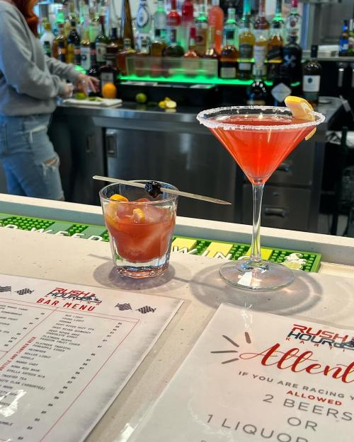 Two colorful cocktails on a bar counter, with a menu and various bottles of liquor in the background.