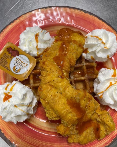 A plate featuring a crispy fried fish fillet on a waffle, topped with syrup and surrounded by whipped cream.