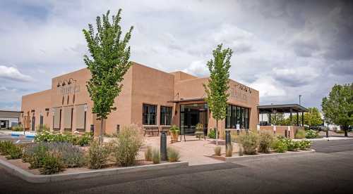 A modern adobe-style building with large windows, surrounded by greenery and a parking lot under a cloudy sky.