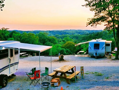 A scenic campsite with RVs, picnic tables, and chairs, overlooking a lush green landscape at sunset.