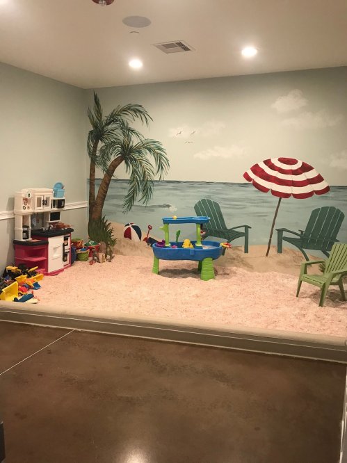 A colorful playroom with a beach mural, toy kitchen, and play area featuring a water table and beach-themed decor.