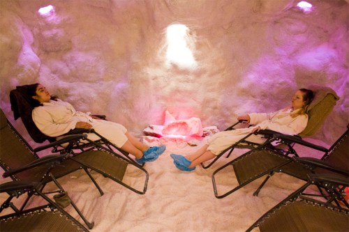 Two people in robes relax in a salt cave, reclining on chairs with a glowing salt lamp in the center.