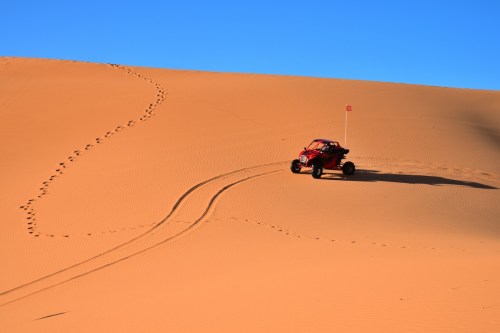 A red off-road vehicle drives on a sandy dune, leaving tire tracks against a clear blue sky.