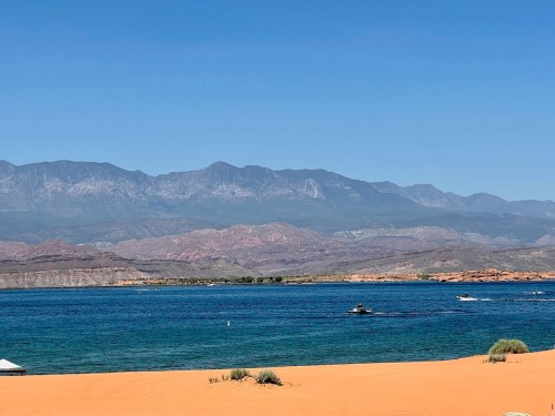 A serene landscape featuring a blue lake surrounded by mountains and sandy shores under a clear blue sky.