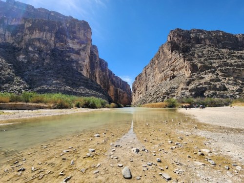 A serene river flows between towering rocky cliffs under a clear blue sky, with greenery along the banks.