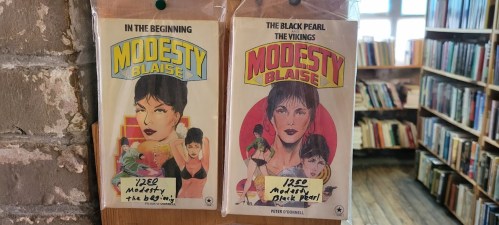 Two Modesty Blaise comic book covers displayed on a shelf in a bookstore, featuring vibrant artwork and titles.
